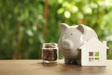 Photo of Piggy bank, house model and coins in glass jar on wooden table outdoors, space for text. Saving money concept