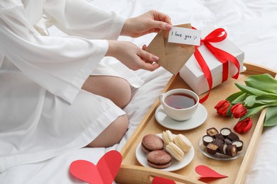 Photo of Tasty breakfast served in bed. Woman with I Love You card, desserts, tea, flowers and gift box at home, closeup