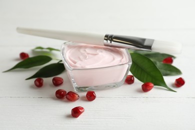 Photo of Glass bowl with natural facial mask, pomegranate seeds and brush on white wooden table, closeup