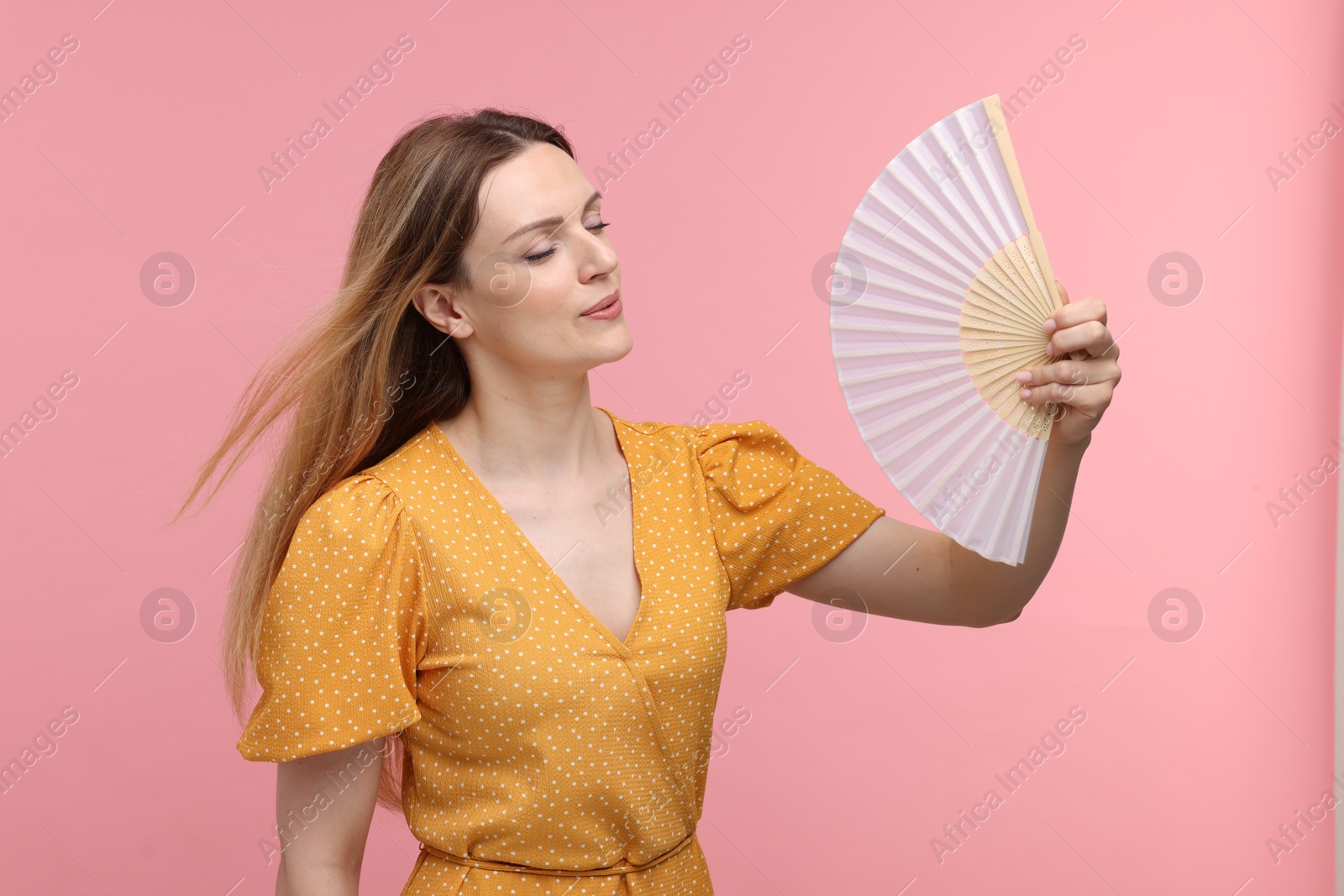 Photo of Beautiful woman waving yellow hand fan to cool herself on pink background