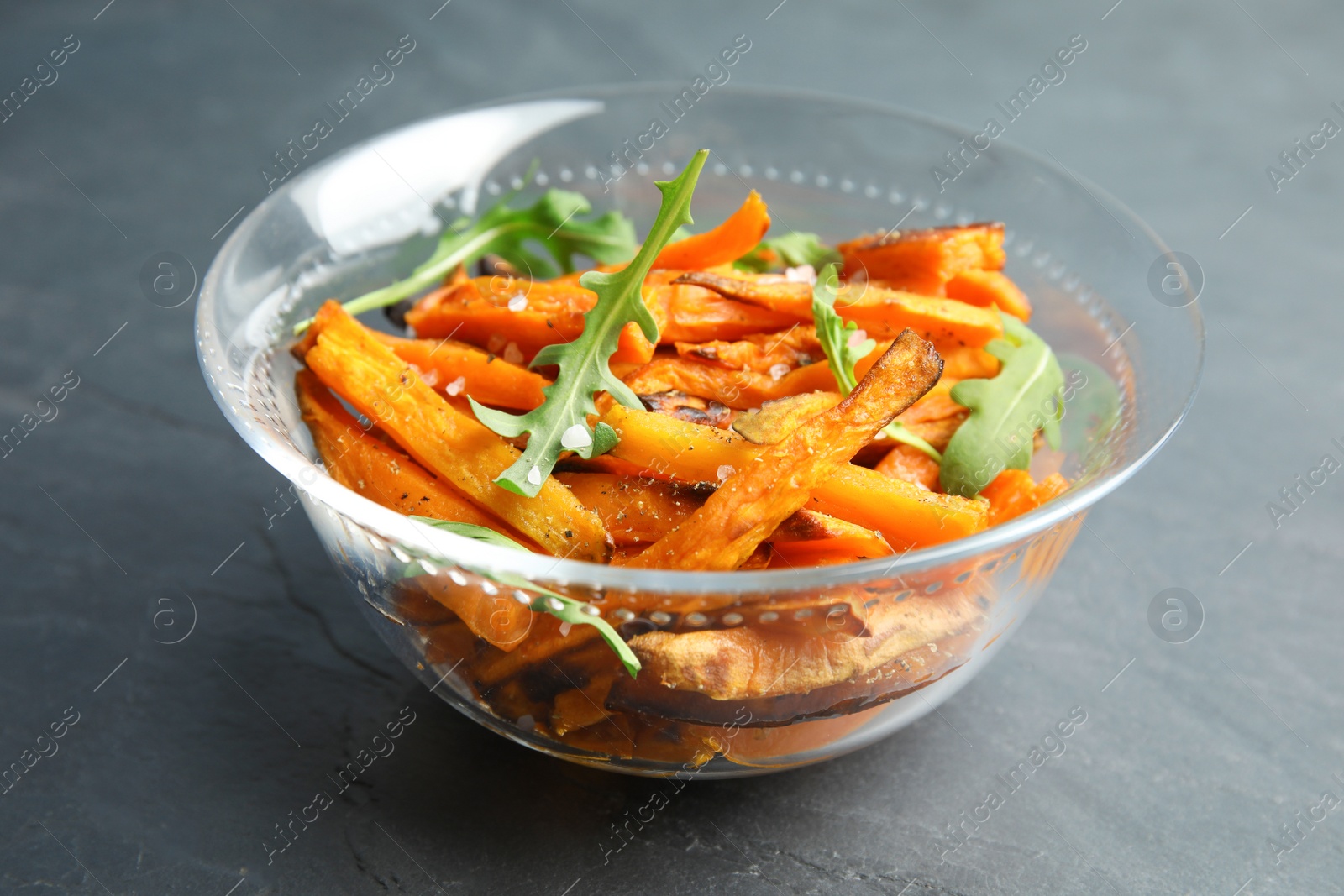 Photo of Glass bowl with baked sweet potato slices and arugula on grey background