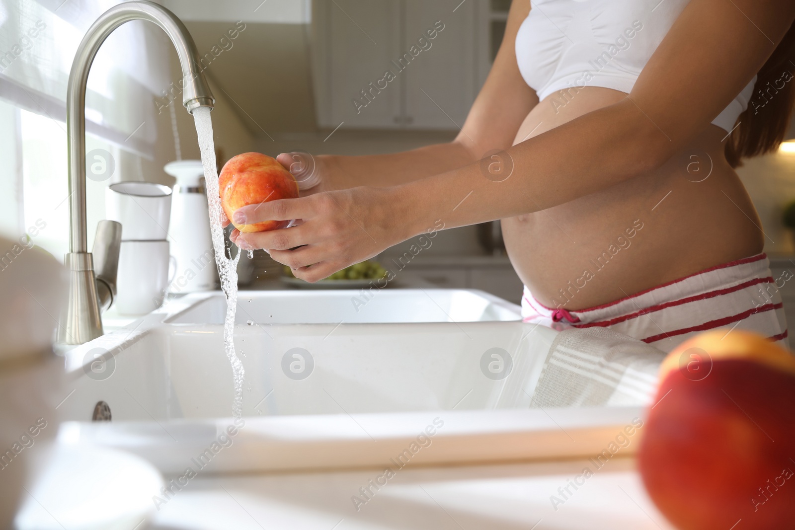 Photo of Young pregnant woman washing fresh sweet peach in kitchen, closeup. Taking care of baby health