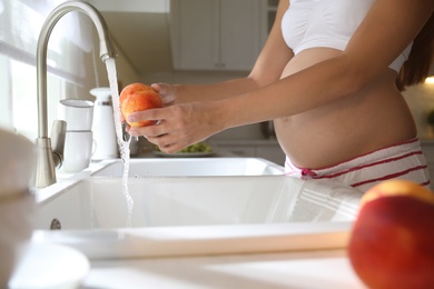 Young pregnant woman washing fresh sweet peach in kitchen, closeup. Taking care of baby health