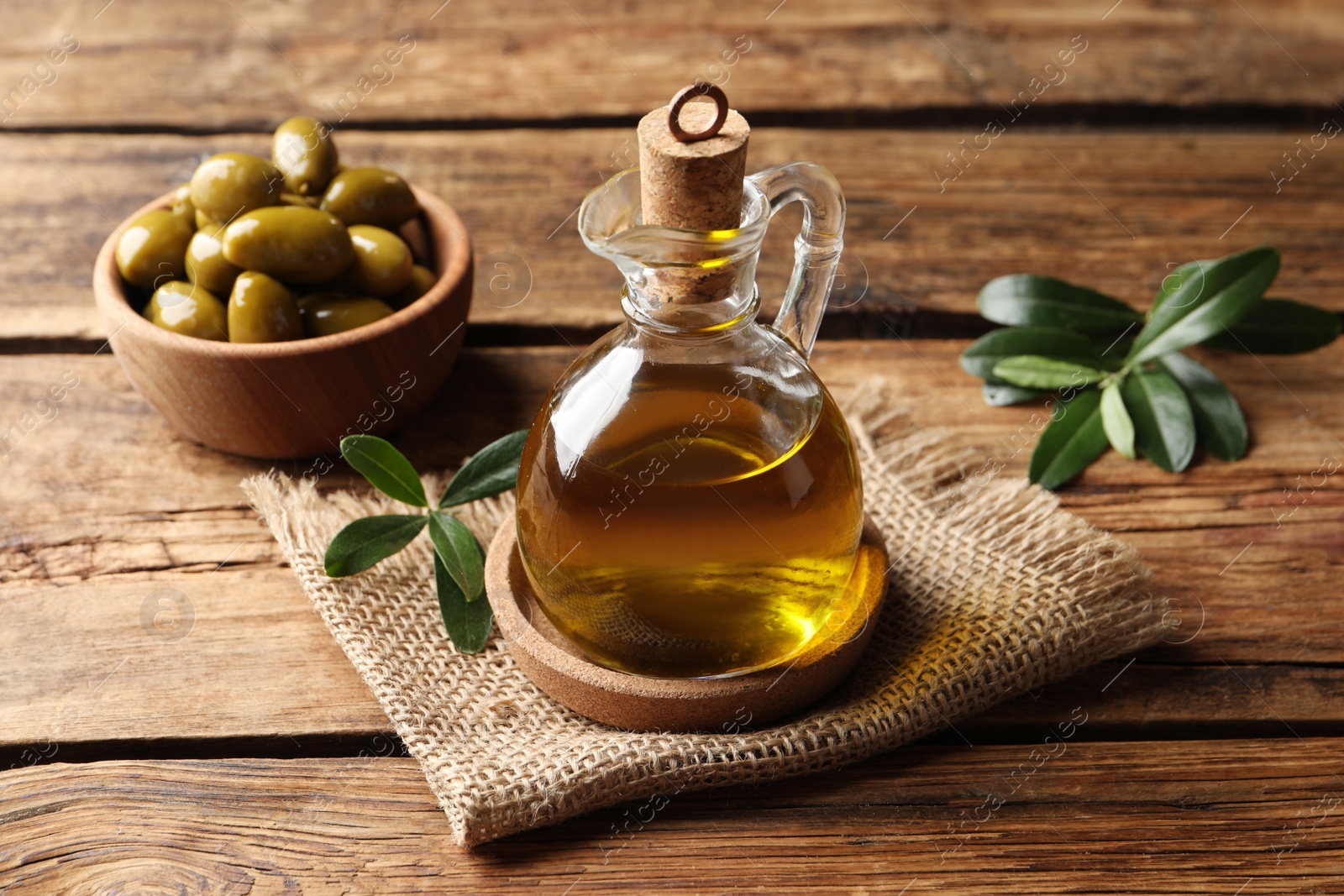 Photo of Glass jug of oil, ripe olives and green leaves on wooden table