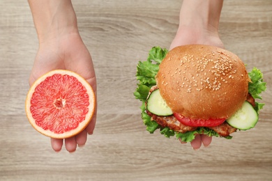 Photo of Woman holding tasty sandwich and half of fresh grapefruit over wooden table, top view. Choice between diet and unhealthy food