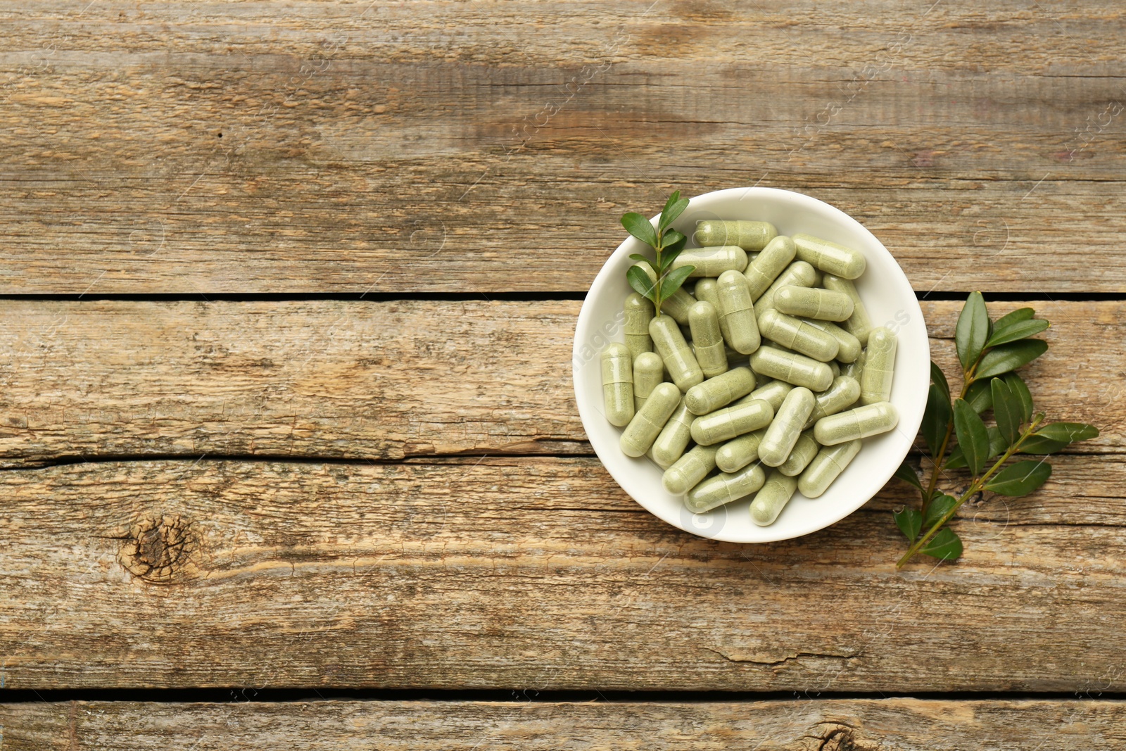 Photo of Vitamin capsules in bowl and leaves on wooden table, top view. Space for text