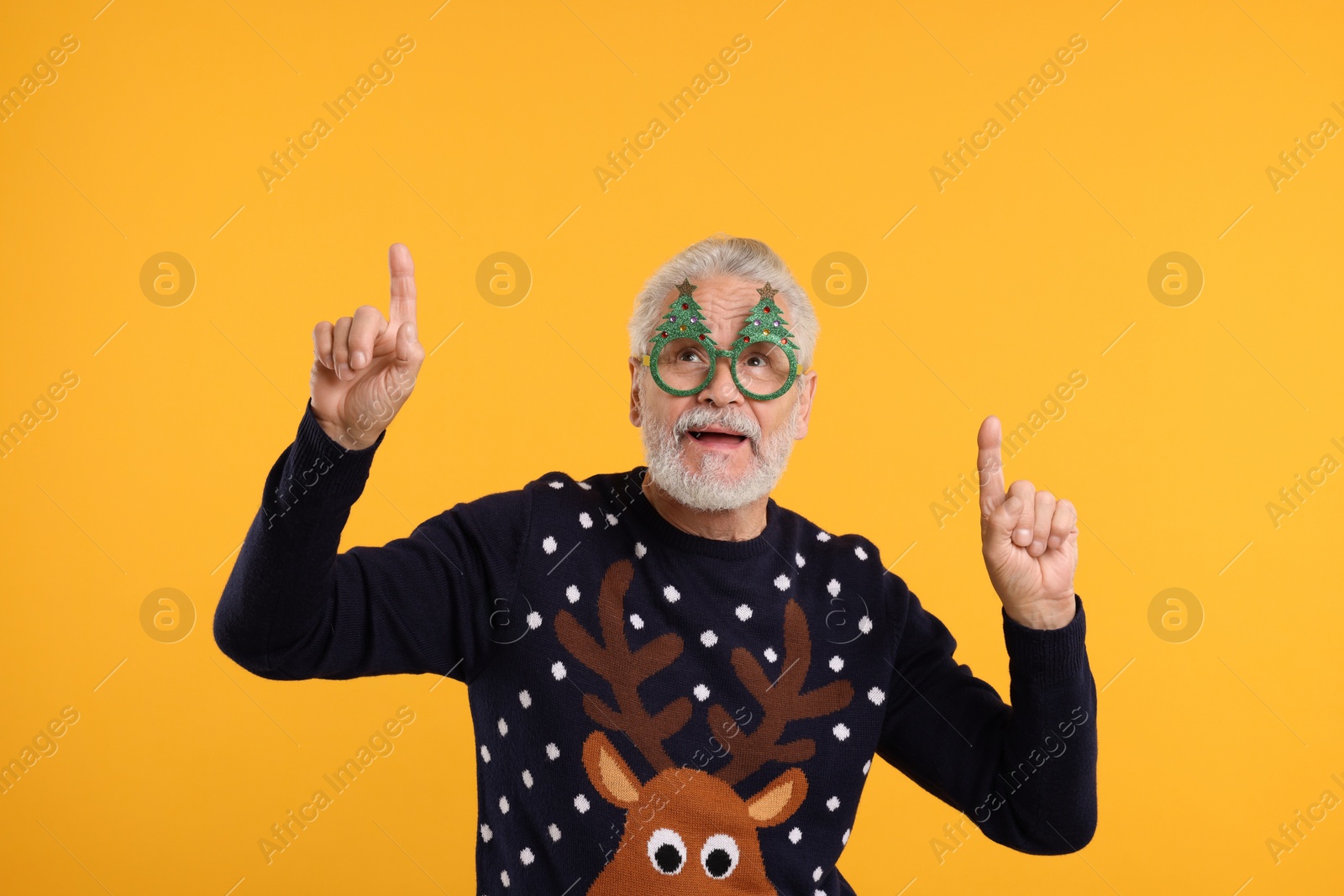 Photo of Senior man in Christmas sweater and funny glasses pointing at something against orange background