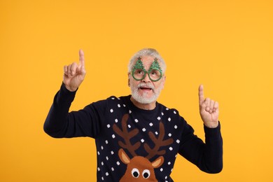 Senior man in Christmas sweater and funny glasses pointing at something against orange background