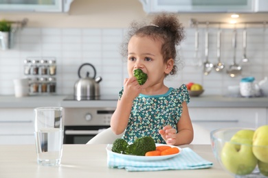 Photo of Cute African-American girl eating vegetables at table in kitchen