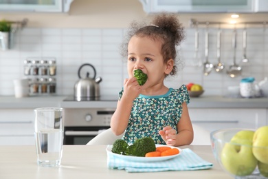 Cute African-American girl eating vegetables at table in kitchen