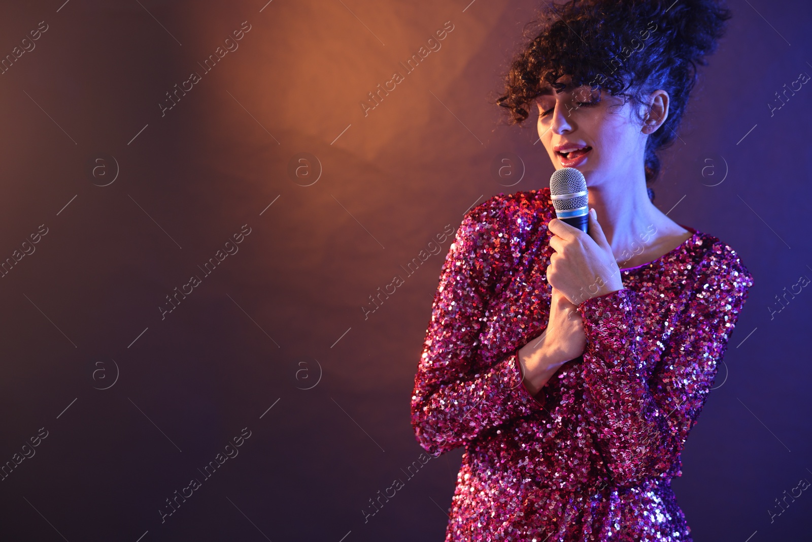 Photo of Beautiful young woman with microphone singing on color background in neon lights. Space for text