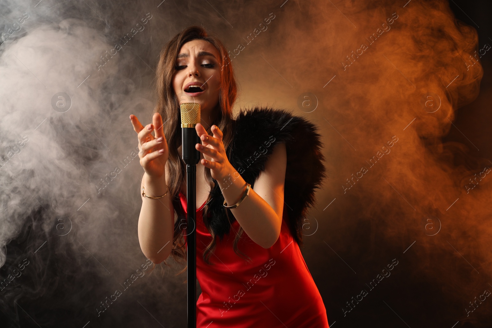 Photo of Beautiful young woman in stylish dress with microphone singing on dark background in color lights and smoke
