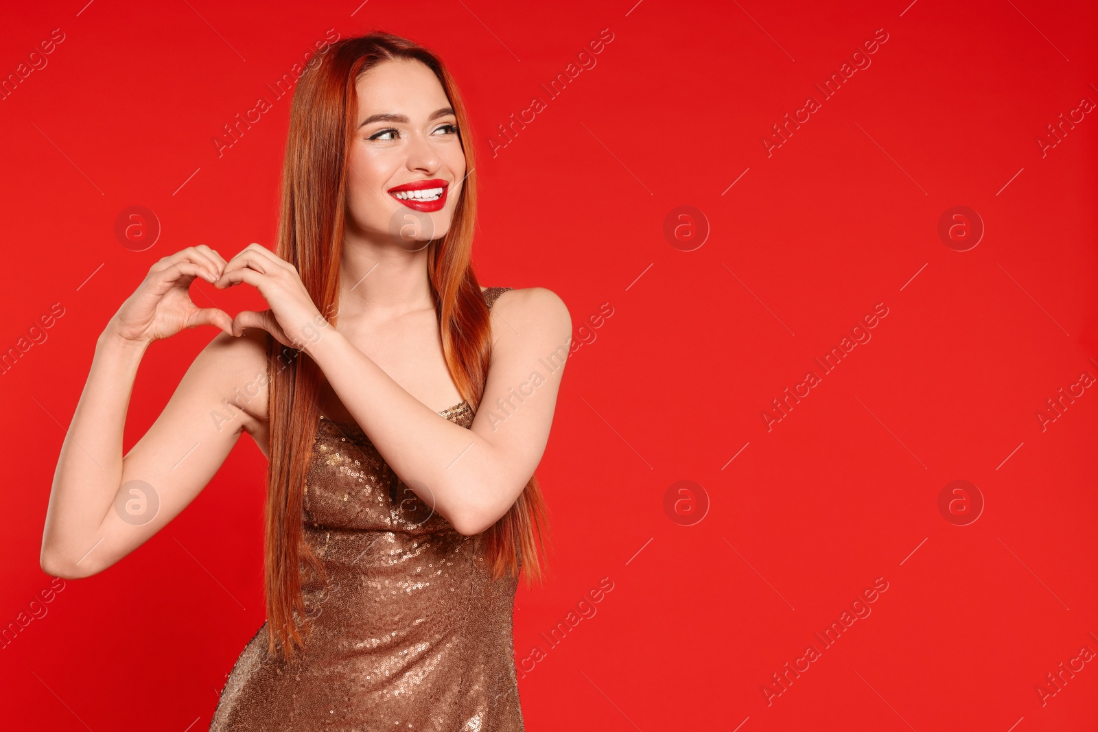 Photo of Young woman in dress making heart with hands on red background, space for text