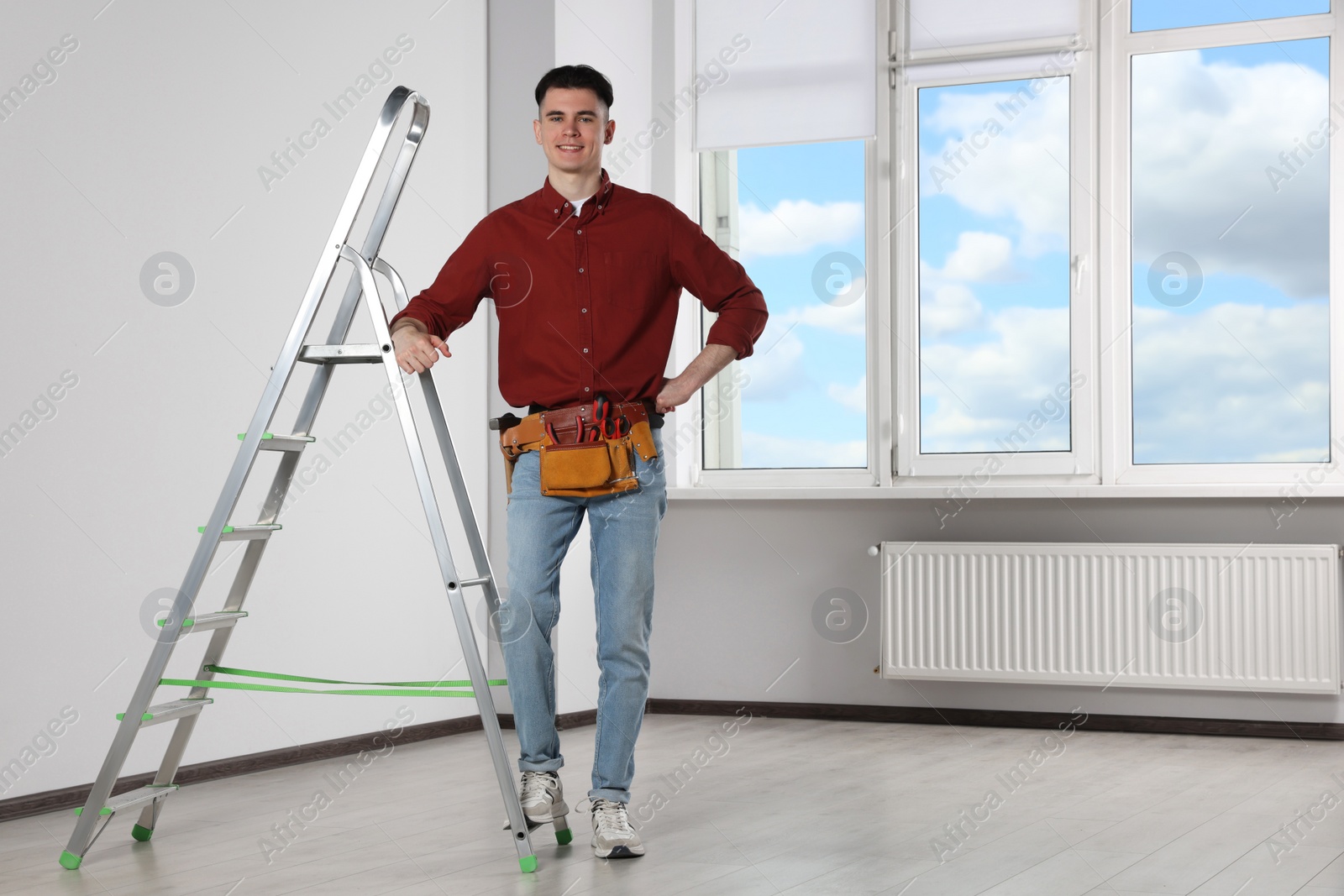Photo of Young handyman with tool belt leaning on stepladder in room