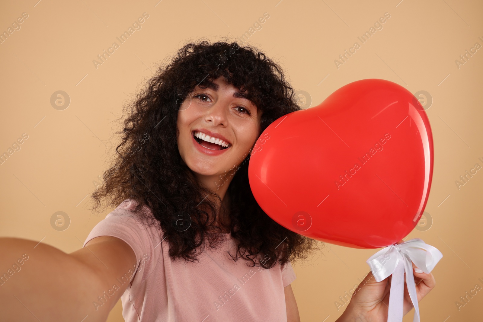 Photo of Happy young woman holding red heart shaped balloon and taking selfie on beige background