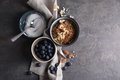 Photo of Tasty granola in bowl, blueberries, yogurt and spoon on gray textured table, flat lay. Space for text