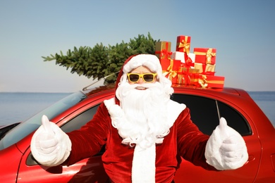 Photo of Authentic Santa Claus near car with presents and fir tree on roof at sea