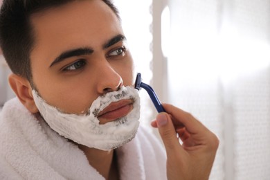 Photo of Handsome young man shaving with razor in bathroom, closeup