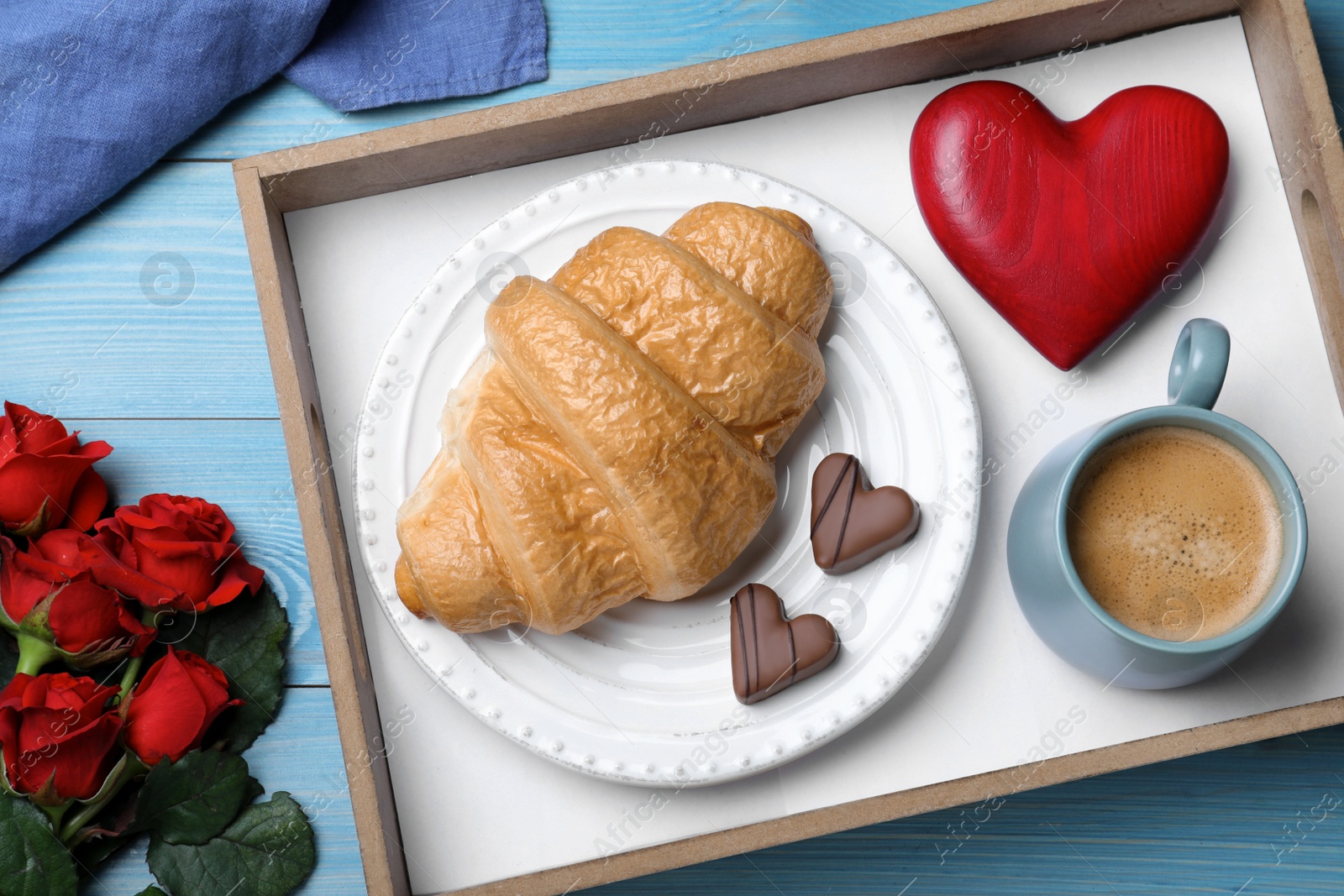 Photo of Romantic breakfast with cup of coffee, croissant and roses on blue wooden table, flat lay. Valentine's day celebration