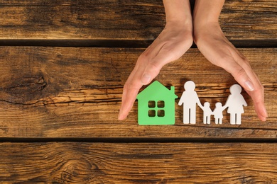 Woman holding hands near figures of house and family on wooden background, top view. Space for text