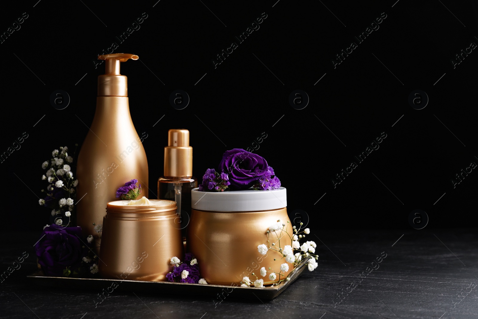 Photo of Set of hair cosmetic products and flowers on stone table against black background. Space for text