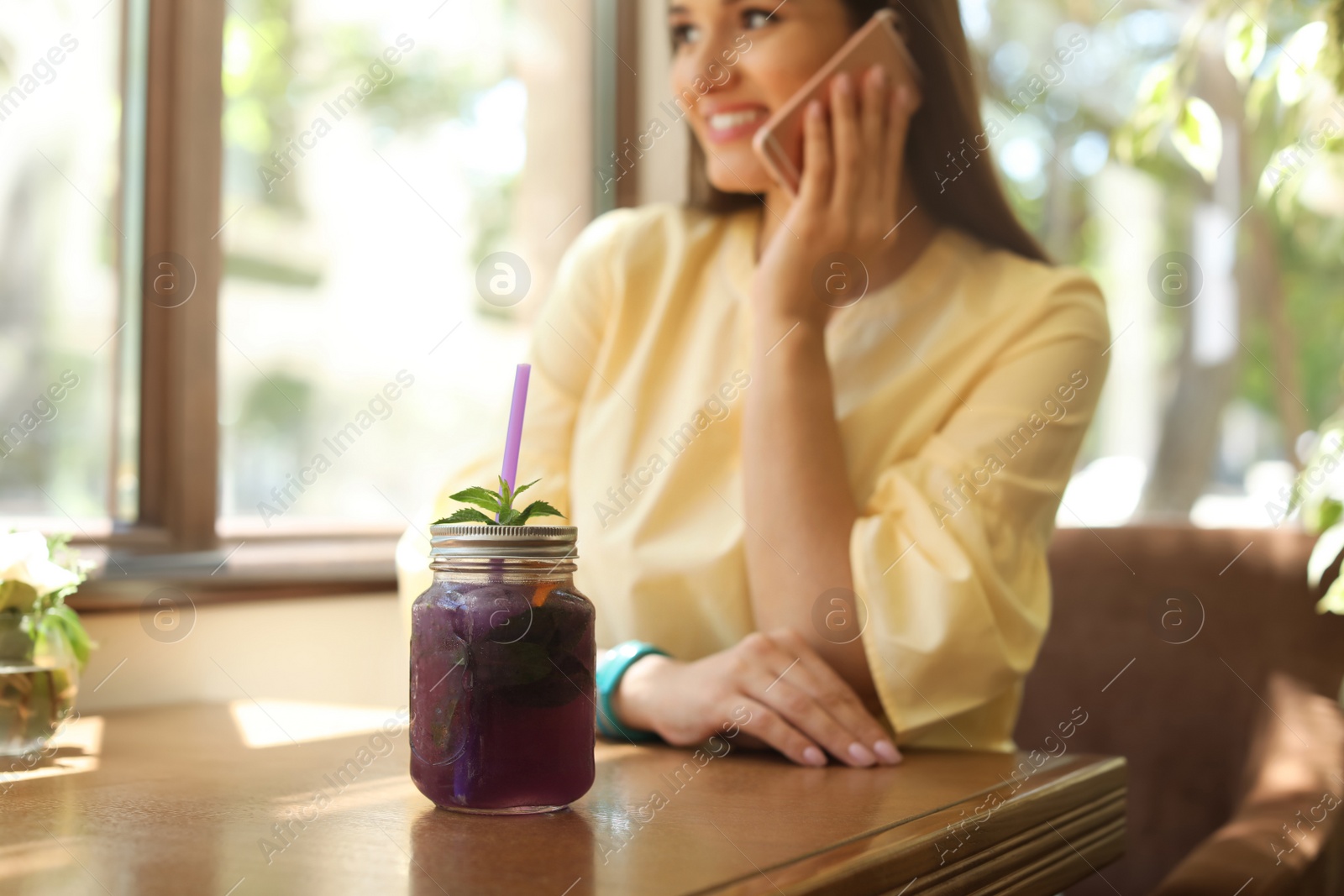 Photo of Mason jar with tasty natural detox lemonade and young woman using mobile phone in cafe