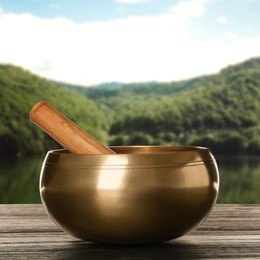 Golden singing bowl and mallet on wooden table against mountain landscape 