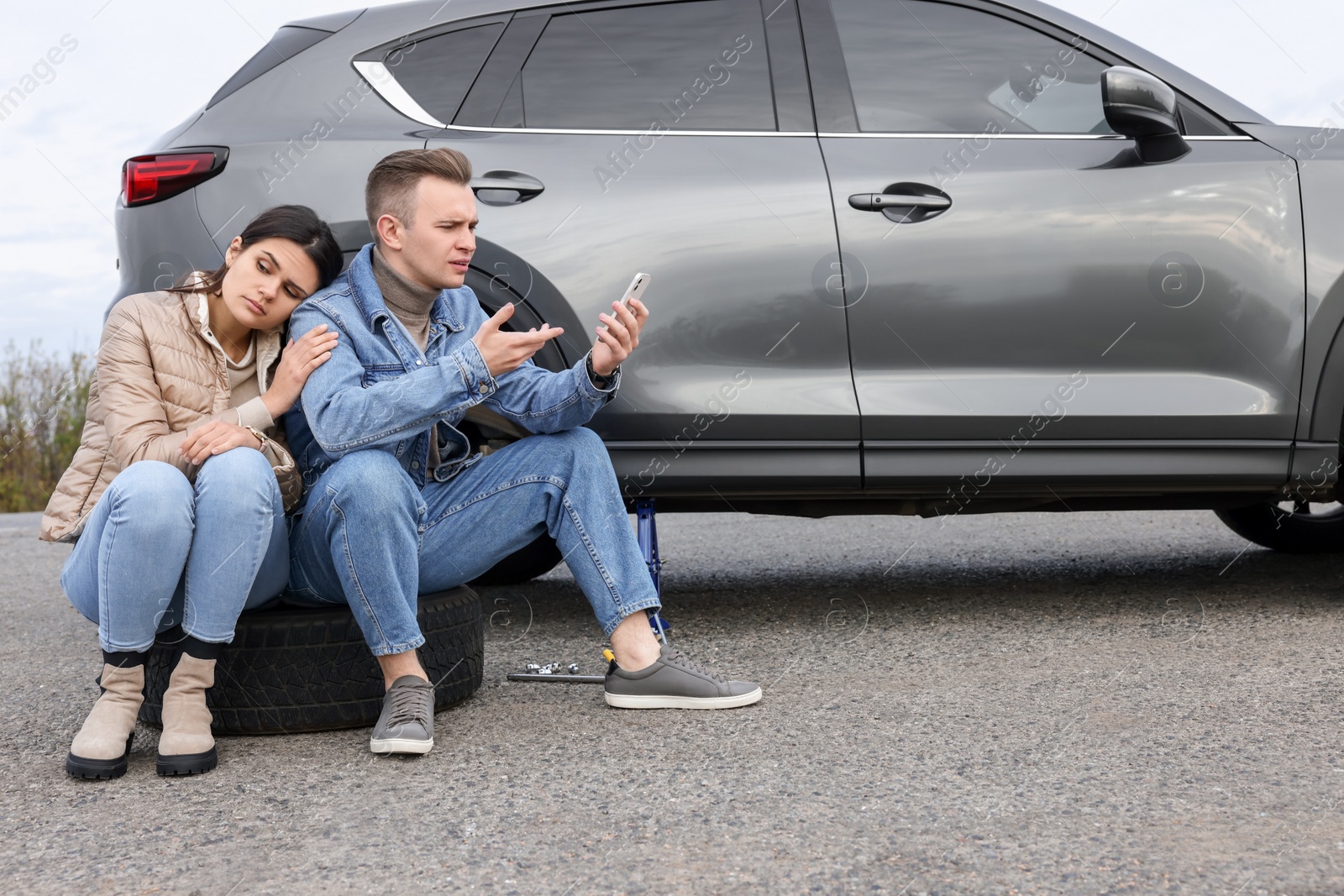 Photo of Young couple with smartphone near car on roadside. Tire puncture