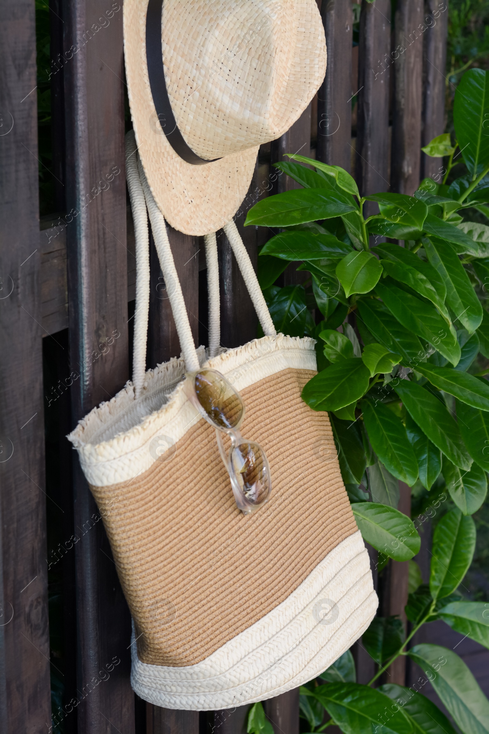 Photo of Stylish bag with hat and sunglasses hanging on wooden fence outdoors. Beach accessories