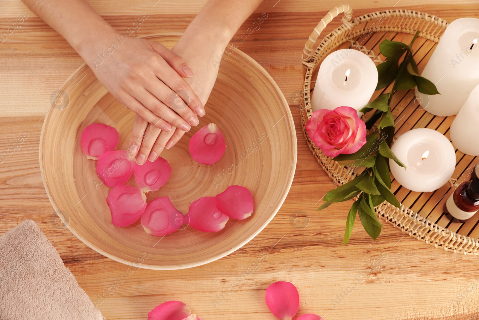 Photo of Woman soaking her hands in bowl with water and petals on wooden table, top view. Spa treatment