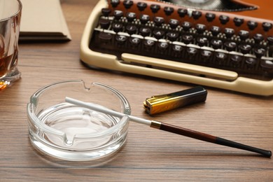 Photo of Glass ashtray with long cigarettes holder and lighter near vintage typewriter on wooden table