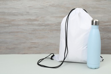 Photo of White drawstring bag and thermo bottle on light table. Space for text