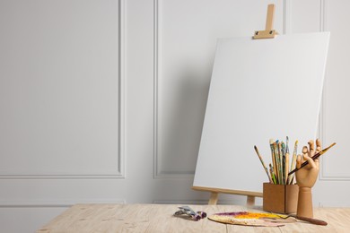 Photo of Easel with blank canvas, hand model and different art supplies on wooden table near white wall. Space for text