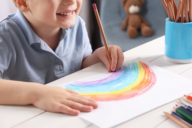 Photo of Little boy drawing rainbow with pencil at white wooden table indoors, closeup. Child`s art
