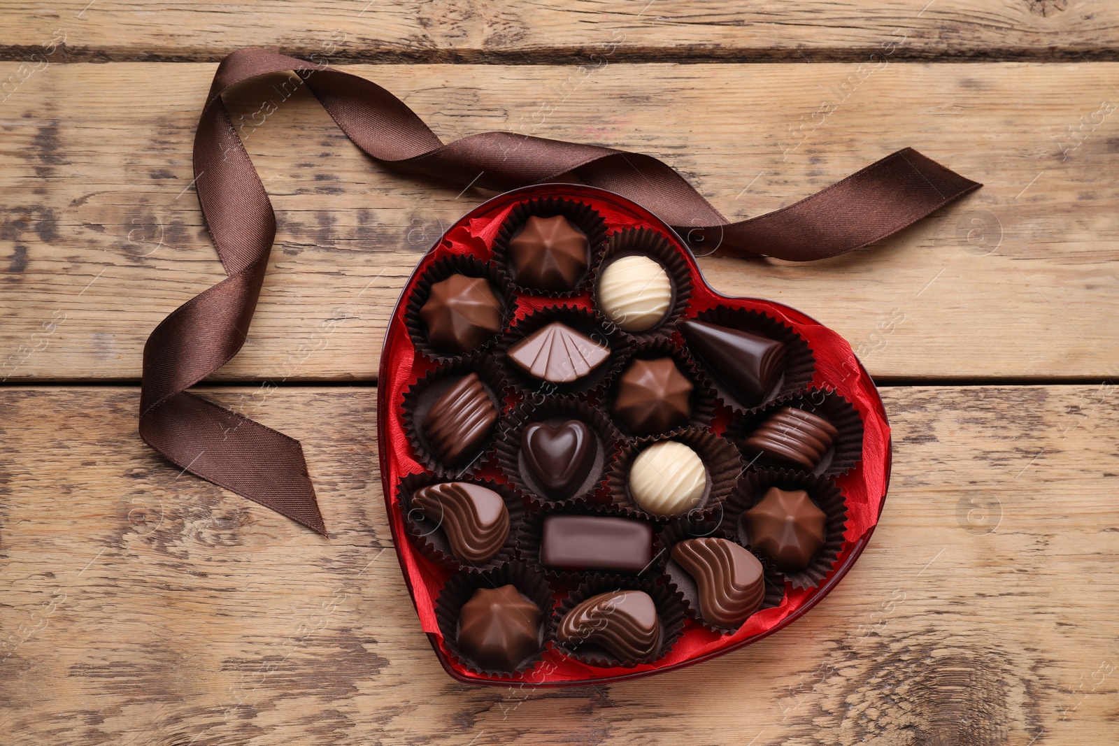 Photo of Heart shaped box with delicious chocolate candies and ribbon on wooden table, flat lay