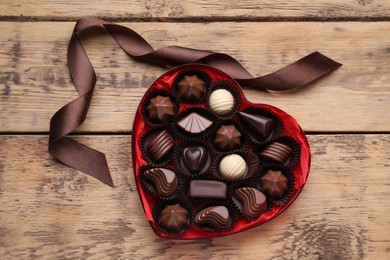 Photo of Heart shaped box with delicious chocolate candies and ribbon on wooden table, flat lay