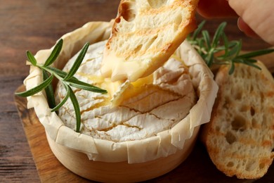 Photo of Dipping bread in tasty baked brie cheese on wooden board, closeup