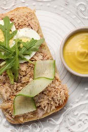 Photo of Delicious sandwich with tuna, boiled egg, vegetables and mustard sauce on white plate, flat lay