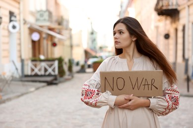 Photo of Sad woman in embroidered dress holding poster No War on city street. Space for text