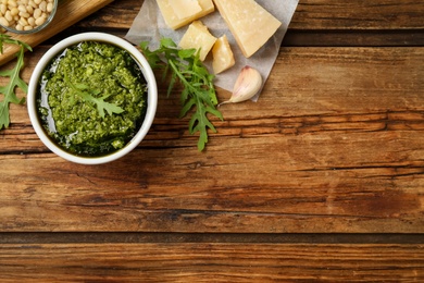 Photo of Bowl of tasty arugula pesto and ingredients on wooden table, flat lay. Space for text