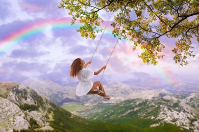 Dream world. Young woman swinging over mountains under sunset sky 