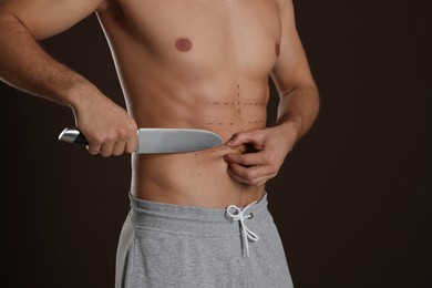 Photo of Fit man with knife and marks on body against dark brown background, closeup. Weight loss surgery