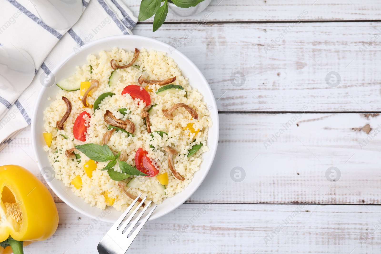 Photo of Bowl of delicious couscous with vegetables and basil served on white wooden table, flat lay. Space for text