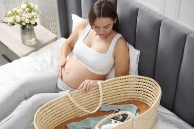 Beautiful pregnant woman with baby basket on bed indoors
