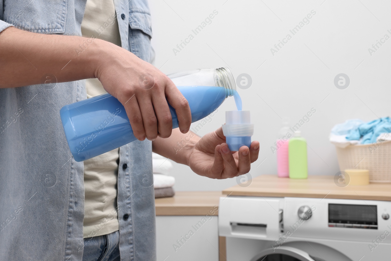 Photo of Man pouring fabric softener from bottle into cap near washing machine indoors, closeup