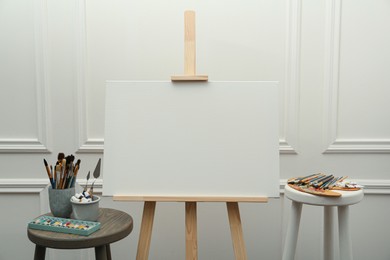 Photo of Easel with blank canvas, brushes, paints and palette in studio