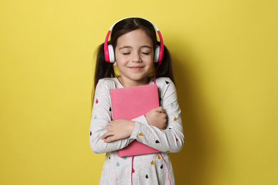 Photo of Cute little girl with headphones listening to audiobook on yellow background