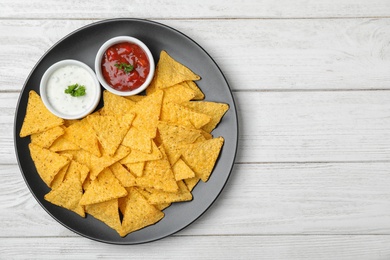 Black plate with tasty Mexican nachos chips and sauces on white wooden table, top view. Space for text