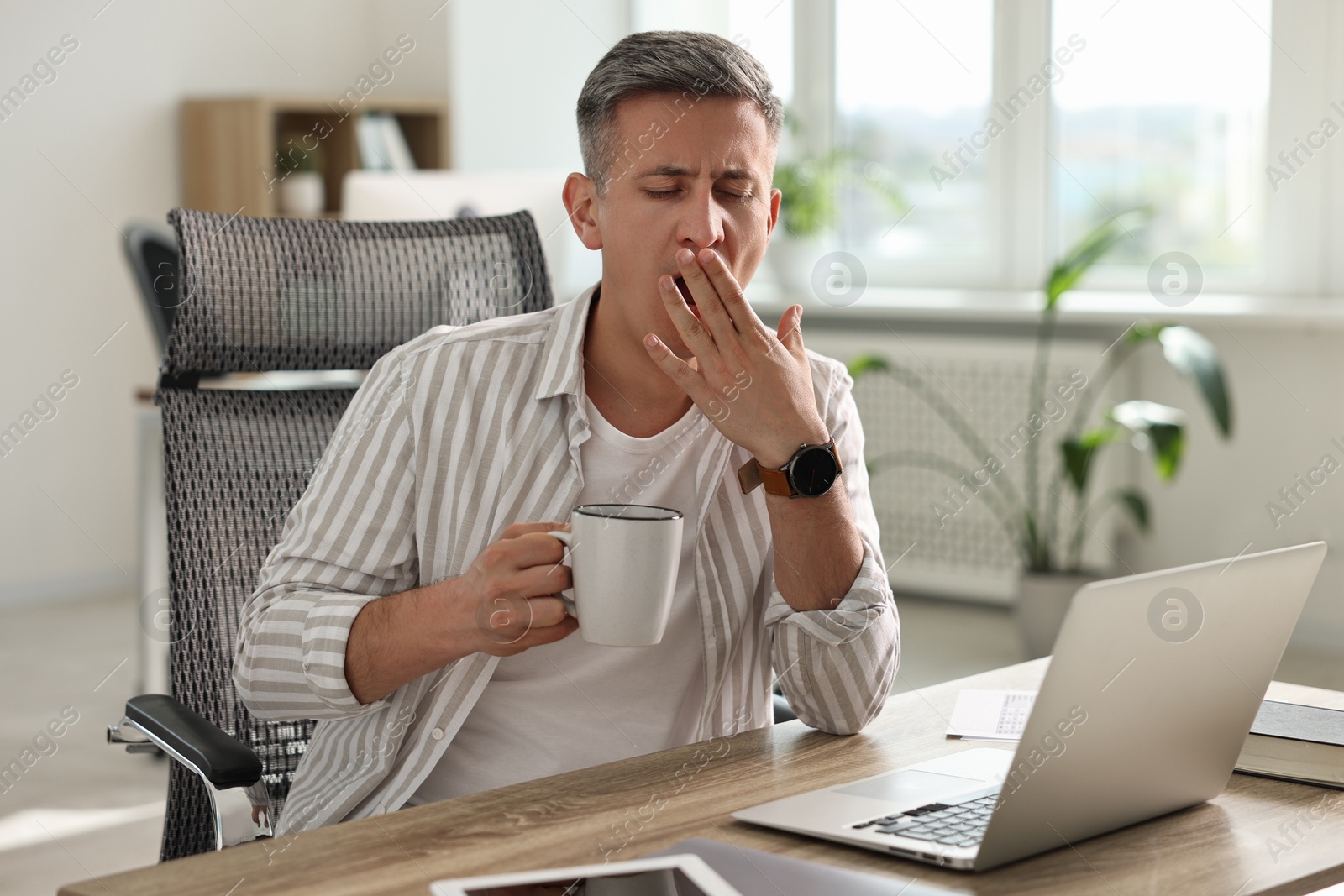 Photo of Man with cup of drink yawning at wooden table in office