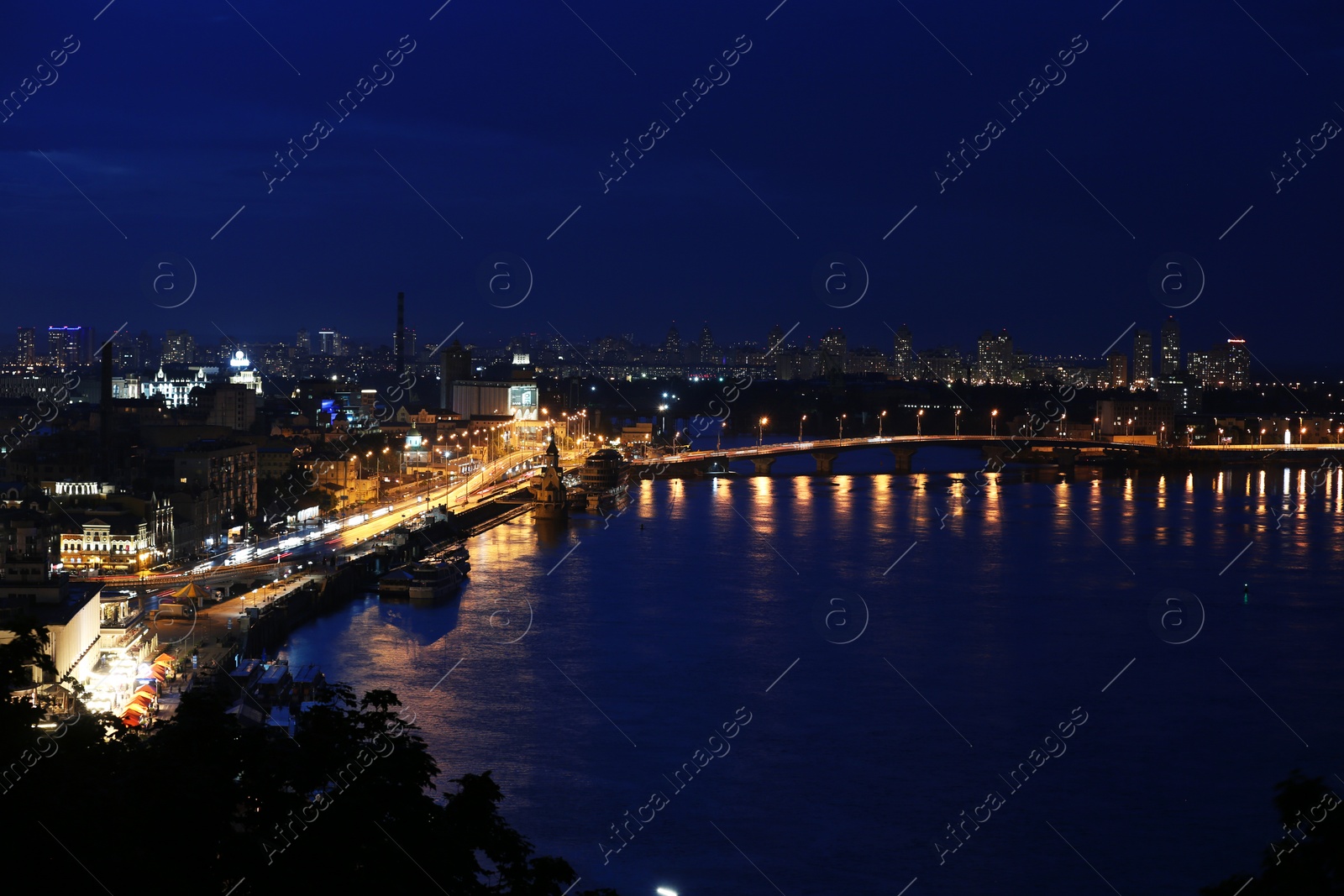 Photo of KYIV, UKRAINE - MAY 21, 2019: Beautiful view of night cityscape with illuminated buildings near river and bridge